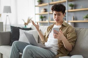 Confused asian man looking at phone screen, sitting on couch with computer on lap in living room, upset frustrated male reading bad news on message, having problem with device or Internet connection photo