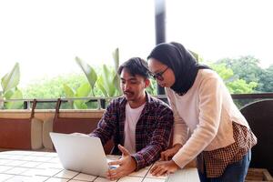 Two Young Asian Working People, Discussing at the Cafe Table, Working with Laptop Explaining File on Screen photo