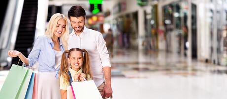 A happy family of three strolls through a shopping mall, with the mother carrying shopping bags and the daughter holding onto her own bags, panorama with copy space photo