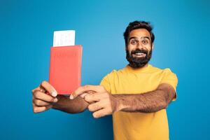 A smiling Indian man wearing a yellow shirt is holding up a red passport with a boarding pass inserted, standing against a bright blue background. photo