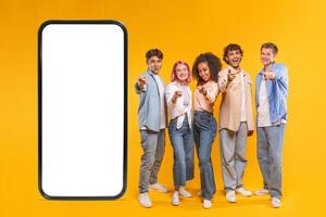 A group of five diverse young adults, all dressed in casual attire, stand in front of a large smartphone with a blank screen. They all point at the screen with a sense of excitement and anticipation. photo