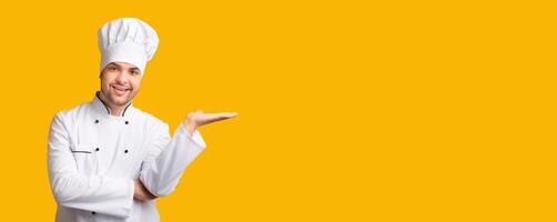 Smiling Chef Man Holding Invisible Object On Open Hand Palm Standing Over Yellow Studio Background. Panorama, Copy Space photo