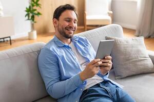 A man is seated on a couch, engrossed in using a tablet computer. He is focused on the screen, tapping and scrolling through content. The room is well-lit photo