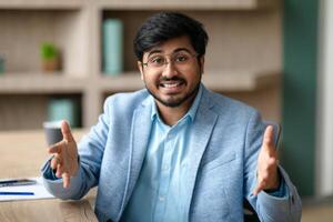 Indian man in a blue blazer and white shirt sits at a desk in an office, smiling and gesturing with both hands. He is looking directly at the camera with a friendly expression. photo