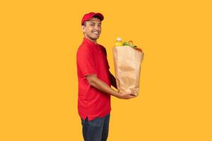 Delivery man standing over yellow studio background, holding a paper bag filled with various food items. He is looking directly at the camera photo