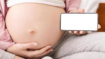 A pregnant woman is depicted holding a smart phone with white screen in her hands, perhaps engaged in messaging, browsing, or other phone-related activities, cropped photo