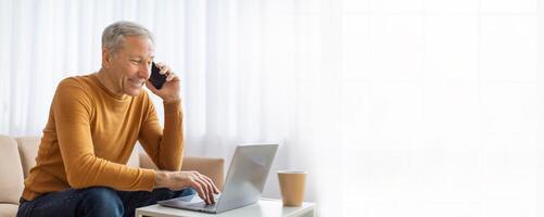 A mature man in a yellow sweater sits on a couch in a bright room while working on a laptop. He is talking on a cell phone and looks at the screen, copy space photo