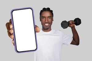 A cheerful young African American man with a bright smile is presenting a smartphone with a blank screen towards the camera, implying an app promotion or tutorial photo