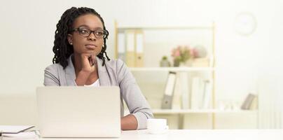 Dreamy black woman is in an office setting using a laptop. The environment is professional and organized, with modern decor and a tidy desk, indicating focus on work, copy space photo