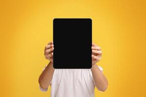 Tablet face. Little boy hiding his face behind portable computer device on orange background, free space photo