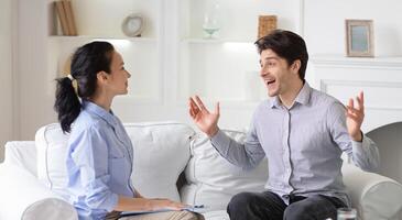 A man animatedly talks with a female therapist in a comfortable living room. Both are seated, engaged in a lively discussion, surrounded by light decor and soft furnishings. photo