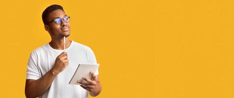 Pensive Redactor. Dreaming afro man in glasses taking notes, writing ideas, looking at copyspace, orange wall, panorama photo