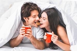 A couple cuddles in bed, sharing a cookie and coffee. They are both smiling and look happy to be together. They are under the covers of a white duvet, with only their heads and shoulders visible. photo
