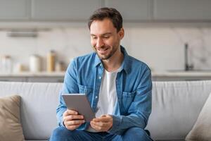 A man in a blue shirt and jeans sits on a light gray couch in a living room. He is looking down at a tablet, smiling, and appears to be browsing the internet or using an app. photo