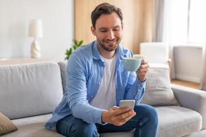 un hombre es sentado en un sofá, participación un taza de café en uno mano y mirando a su teléfono en el otro. él aparece relajado y enfocado en su teléfono pantalla. foto