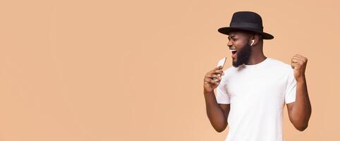 African American man with a beard and a black hat is singing into his phone. His right hand is raised in the air, and he is smiling and looking at copy space photo