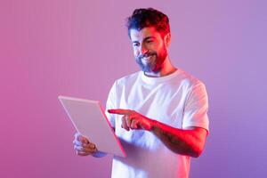 A bearded man wearing a white t-shirt interacts with a digital tablet while standing in a studio with purple and pink neon lighting. He is smiling and appears engaged with the content on the tablet. photo
