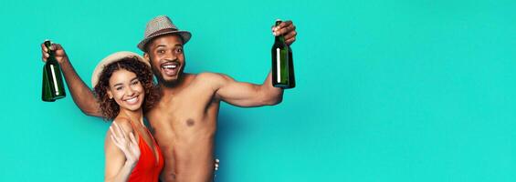 African American couple is smiling and holding bottles in front of a bright turquoise background. The man is shirtless and wearing a hat, and the woman is wearing a red swimsuit and a straw hat. photo