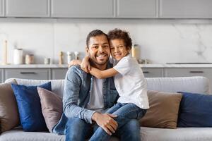 African American man is sitting on top of a couch next to a little boy. The father appears relaxed, while the son looks curious. They seem to be engaged in a conversation or activity together. photo