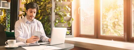 A man sits at a desk using a smartphone while working on a laptop, surrounded by plants and sunlight streaming through the window, copy space photo