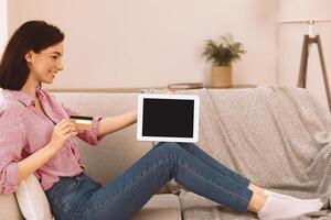 Buy With Our App And Website. Happy girl holding credit card and showing blank tablet screen, sitting on couch, mock up photo