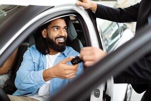 A cheerful Indian man is seated inside a car, receiving a set of car keys from a salesperson, suggesting he has possibly made a purchase or is taking a test drive photo