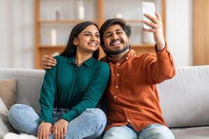 Indian man and woman are sitting side by side on a gray couch, smiling at the camera as they take a selfie together. The man is holding the phone out in front of them, capturing the moment. photo