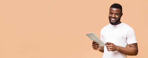 A young black man with a beard smiles while holding a tablet and looking at the camera. He is wearing a white t-shirt. The background is a peach color, copy space photo