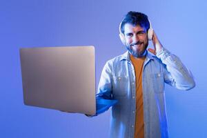 A cheerful young man is holding a laptop in one hand while listening to music with headphones. Background is illuminated with soft blue lighting, indicating he is likely enjoying a nighttime session photo