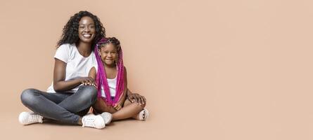Beautiful african american mother sitting on floor with her cute little child, and smiling to camera, yellow studio background with empty space photo