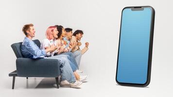 Multiracial group of young people sitting on couch, looking at huge phone with blank blue screen, isolated on white studio background, mockup, copy space photo