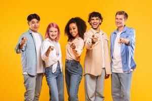 A diverse group of five young adults are standing in front of a yellow background, all pointing at the camera with big smiles on their faces. They are all dressed casually in jeans and shirts. photo