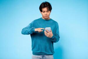 Asian guy wearing a blue sweater is focused on his cell phone screen, tapping and scrolling with his fingers. He appears engrossed in whatever content is displayed on his device. photo