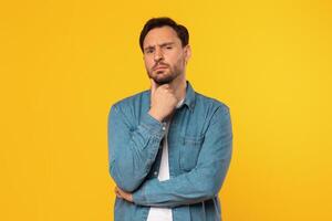 Man with a beard, wearing a blue denim shirt, stands in front of a yellow background, looking directly at the camera with a thoughtful expression. He has his hand on his chin and his arms are crossed photo