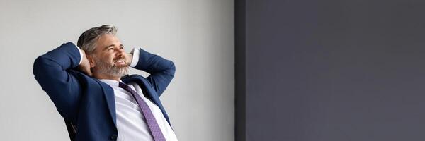 Smiling middle aged businessman leaning back in chair at workplace in office, handsome happy entrepreneur in suit sitting at desk with hands behind head, resting after successful day, copy space photo