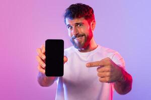 A bearded man wearing a white t-shirt is smiling and pointing at a smartphone with empty screen he is holding, against a vibrant purple and pink gradient background. photo