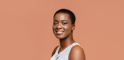 A portrait of a smiling Black woman with short hair. She is wearing a white and black striped tank top and is looking at the camera. The background is a peach color. photo