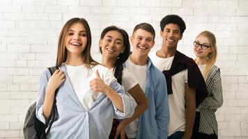 A diverse group of multiethnic teenagers is lined up, displaying a variety of cheerful expressions. The backdrop features a white brick wall, suggesting an informal or educational setting photo