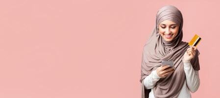 Online banking. Happy arabic girl in hijab using smartphone and holding credit card, enjoying easy payments or internet shopping, pink background photo