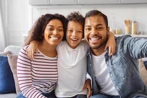 Black family consisting of parents and child are sitting on a couch, holding a smartphone in front of them to take a selfie together. They are smiling and posing for the camera photo