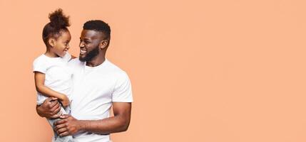 An African American father smiles and holds his little daughter in his arms against a peach-colored background. They are both looking at each other and laughing, copy space photo