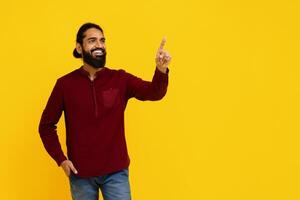 Indian cheerful man with a beard is standing against a bright yellow backdrop, pointing upwards with his left hand, touching invisible virtual screen photo