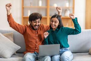 Indian man and woman are seated side by side on a couch, focusing intently on a laptop screen. They appear engaged in an online activity or work-related task, with the laptop placed on their laps photo