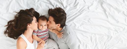 A young couple is tenderly kissing each other while holding their adorable baby in the center. They are lying on a soft white bed, radiating warmth and affection, top view, copy space photo