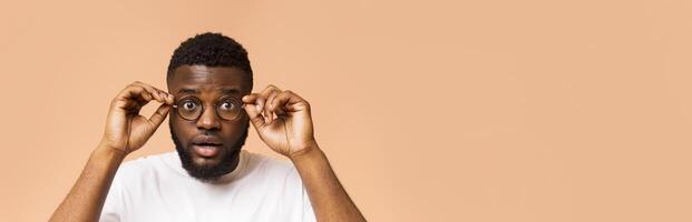 This image features a young black man of color wearing a white T-shirt with a surprised expression on his face. He is holding his round eyeglasses in front of his face with both hands, copy space photo