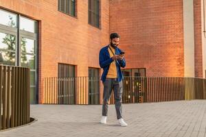 Indian man is standing in front of a brick building, focused on his cell phone screen. He appears engrossed in his device, with the urban backdrop of the building behind him. photo