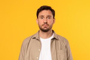 A man with a beard and brown hair is wearing a beige button-up shirt over a white t-shirt. He is looking directly at the camera with a serious expression on his face. The background is a solid yellow. photo