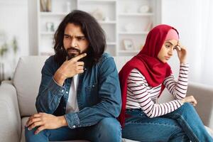A bearded Indian man and a woman in a red hijab are sitting separately on a couch, showing expressions of concern and frustration, suggesting a difficult conversation or disagreement photo