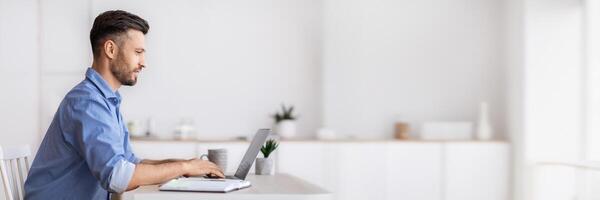 Handsome millennial businessman sitting at workplace in white office, working on laptop, typing on computer keyboard, answering emails, corresponding with clients, side view with copy space photo
