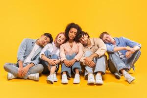 Five multiracial friends are sitting on a yellow background and taking a nap together. They are all wearing casual clothing and have their heads resting on each others shoulders. photo